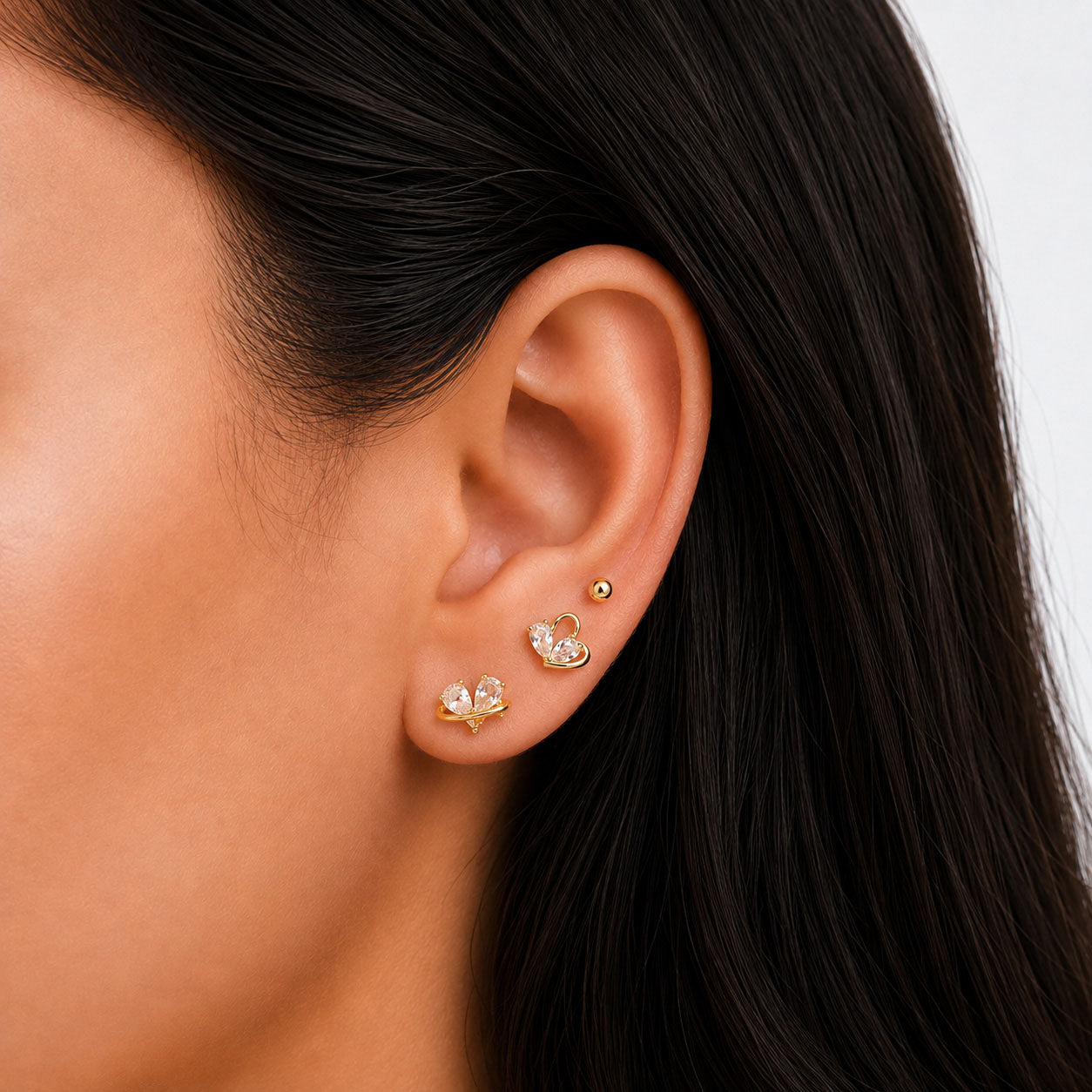 Close-up view of a model’s ear wearing gold-tone butterfly earrings with clear crystal details, paired with small gold ball stud earrings on a clean white background.
