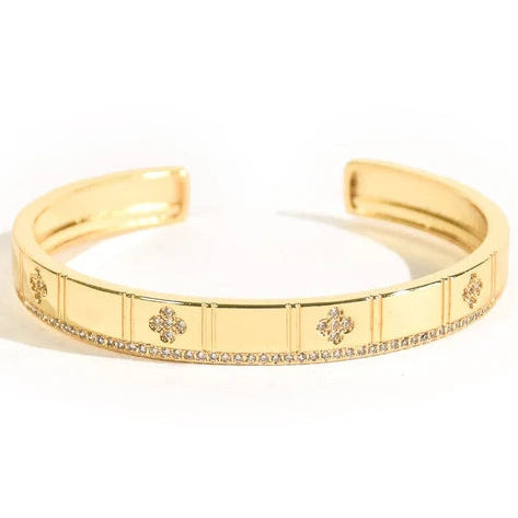 The image shows a gold openable cuff bangle with a sleek rectangular-panel design, small floral/crystal motifs across the front, and a continuous row of pavé-style stones along the lower edge. It is photographed on a clean white background with bright studio lighting and a soft natural shadow.