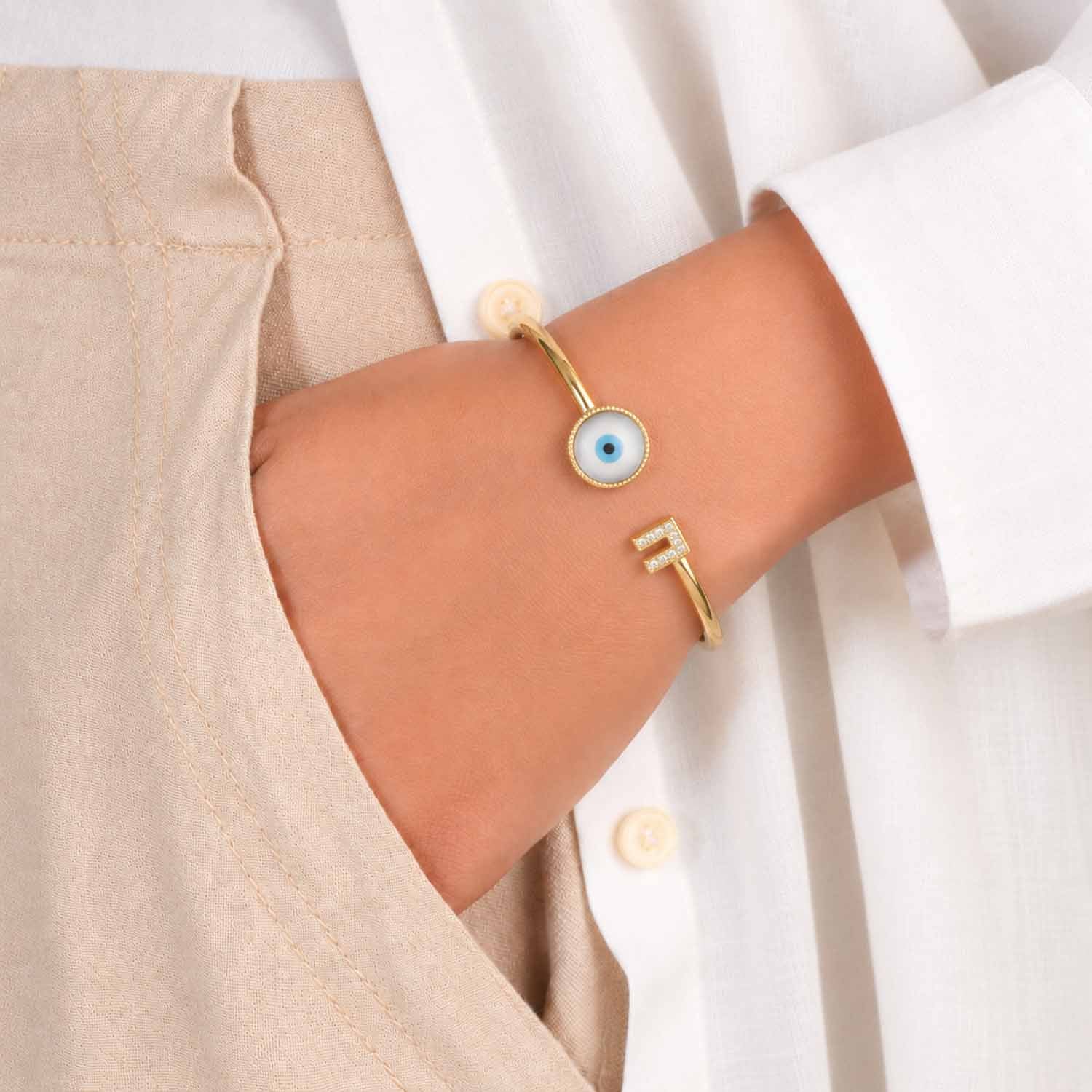Gold open cuff bangle with a blue evil eye centerpiece and crystal-studded geometric detail, worn on the wrist with a neutral beige and white outfit.