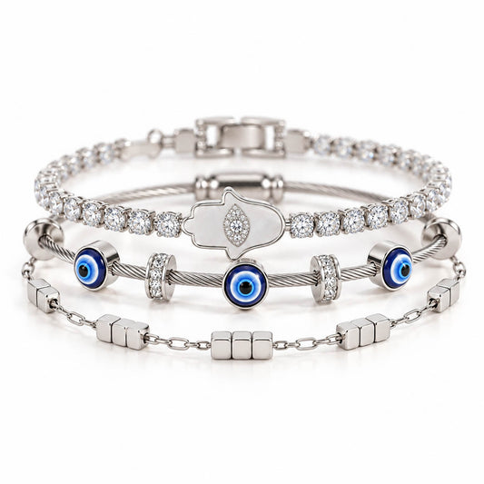 A product photo of a stacked set of silver-tone bracelets featuring a crystal tennis chain with a mother-of-pearl Hamsa hand pendant, a twisted cable wire band with blue evil eye beads, and a link chain with small cube charms.