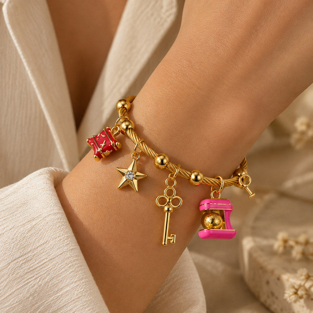 Close-up of a person wearing a gold-tone charm bracelet with a twisted chain and multiple dangling charms, including a red gift box, star, key, and pink capsule-shaped charm, styled with a white outfit against a warm neutral background.