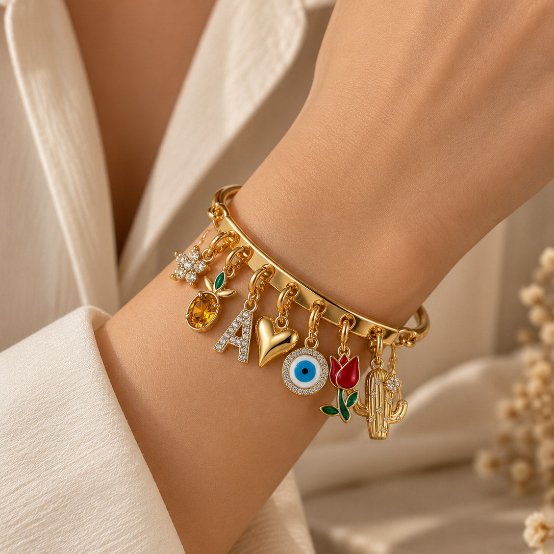 Close-up of a person wearing a gold-tone charm bracelet with multiple dangling charms, including a crystal-studded flower, amber gemstone, letter “A,” heart, blue evil eye, red tulip, and cactus, styled with a white outfit against a warm neutral background.