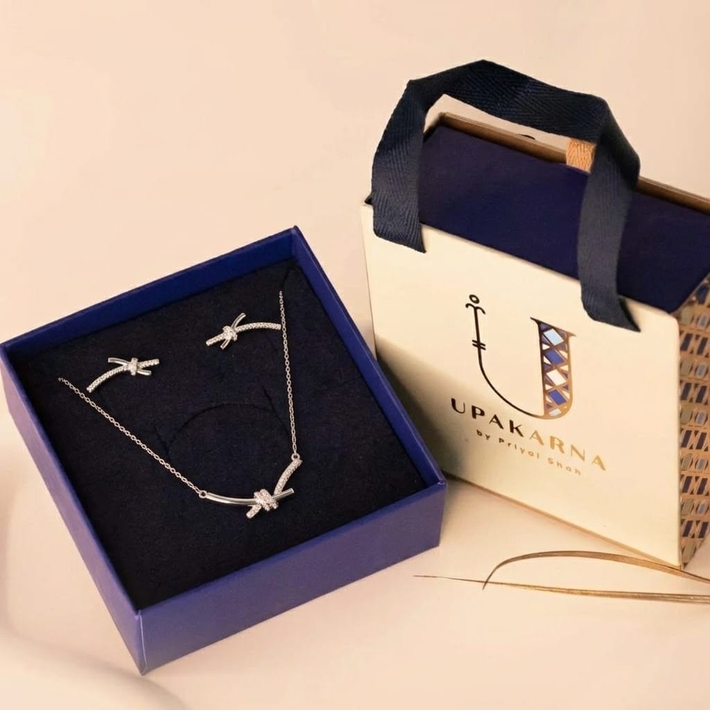 Silver knot-design necklace set with crystal detailing in a navy box.
Paired with an Upakarna gift bag on a beige background.