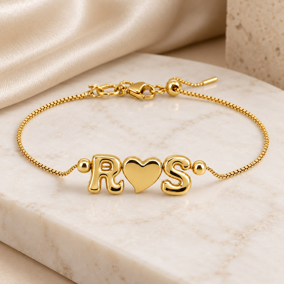 Gold-tone personalized bracelet featuring R♥S letter charms on a delicate adjustable chain, displayed on a cream marble surface with a soft satin background.