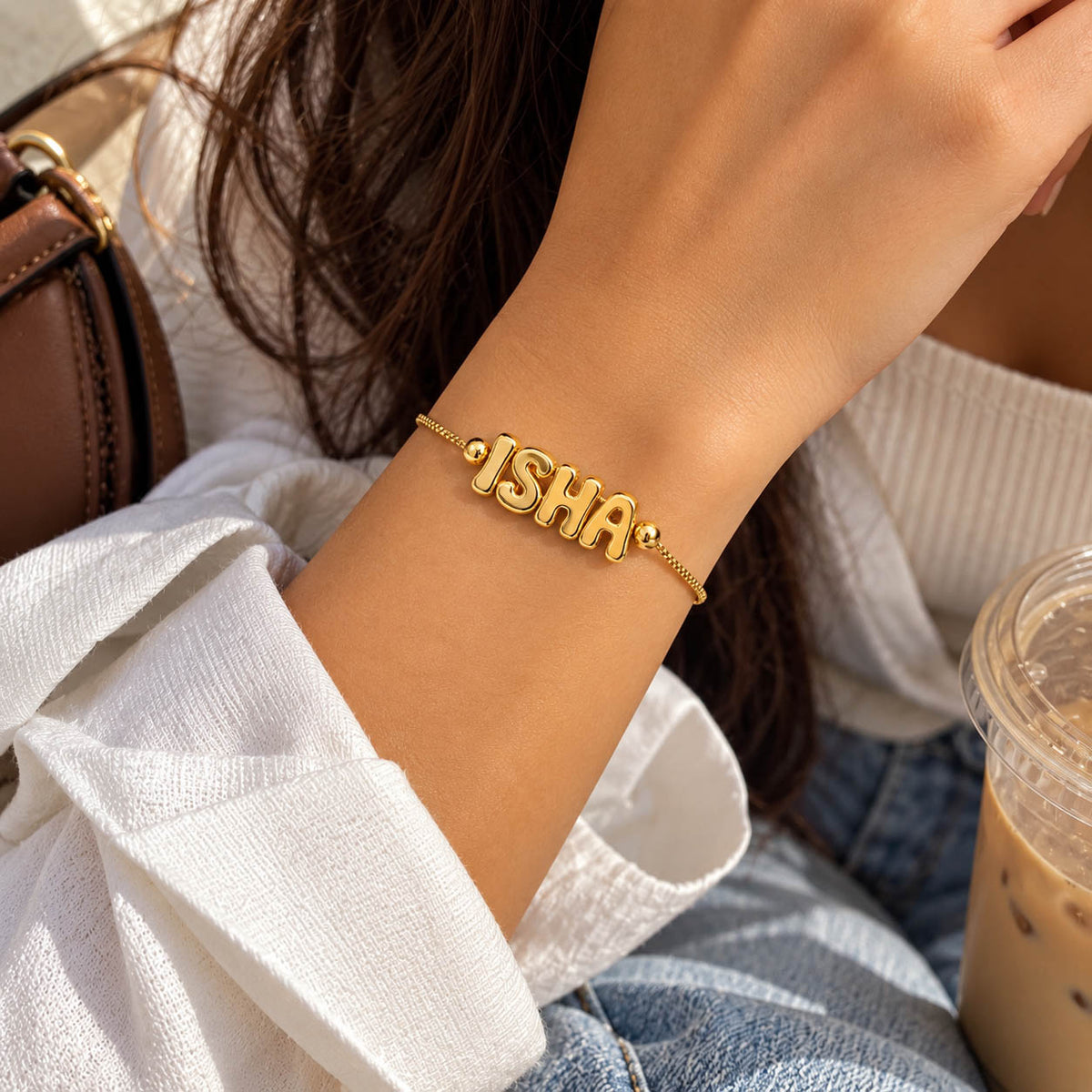 A lifestyle photograph of a person's wrist wearing a gold-toned adjustable box-chain bracelet featuring chunky, puffed bubble-letter charms spelling "ISHA", set against a blurred background with a white sleeve, denim jeans, and a iced coffee cup.