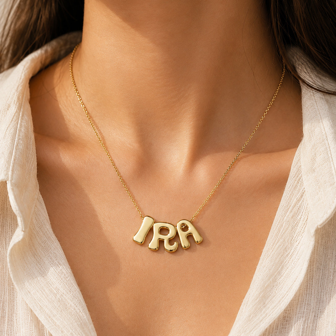 Gold-plated personalized name necklace featuring the name “IRA” on a delicate gold chain, worn on a model in a close-up with a white outfit and warm natural lighting.