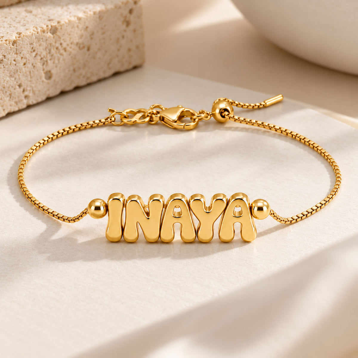 A studio product photograph of a gold-toned adjustable box-chain bracelet featuring chunky, puffed bubble-letter charms spelling "INAYA", laid out cleanly on a textured neutral surface next to a stone block and a white bowl