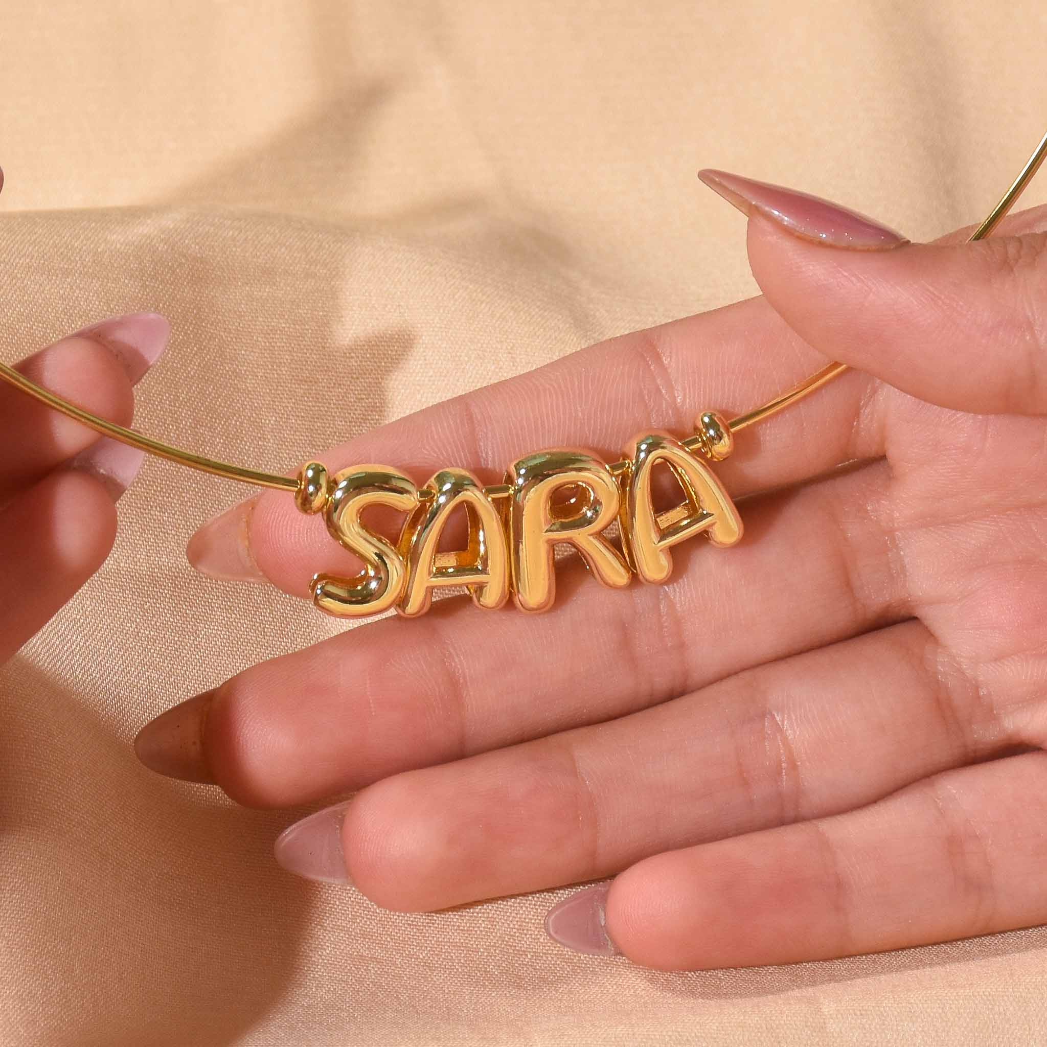 Close-up of a personalised gold-tone necklace featuring the name “SARA” in bold letter charms, held delicately by hands against a soft neutral background.