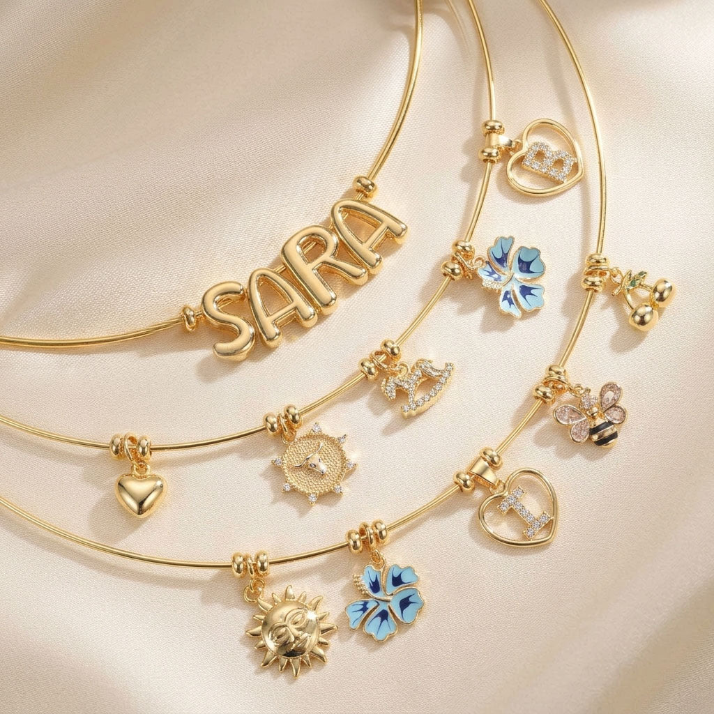 Personalised gold charm necklaces featuring the name SARA, sparkling initial charms, heart charms, blue butterfly charms, sun charms, bee charms, zodiac-style charms, and delicate decorative pendants.