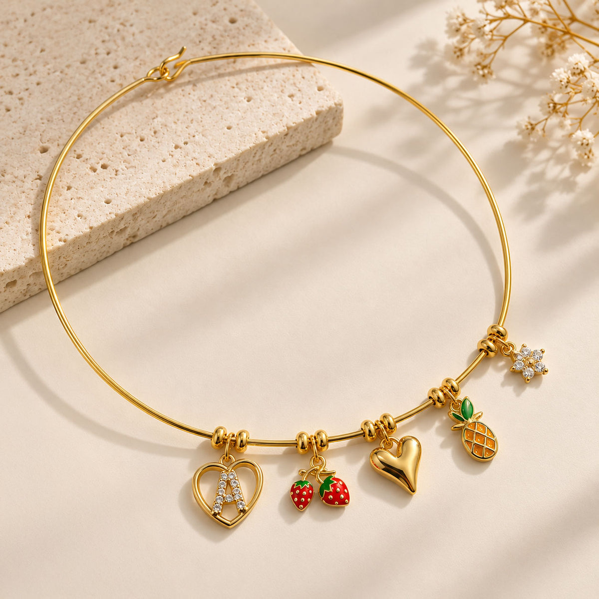Gold tone charm necklace arranged on a beige surface, featuring a heart-shaped initial A charm, strawberry charms, a pineapple charm, and crystal detailing with soft floral accents.
