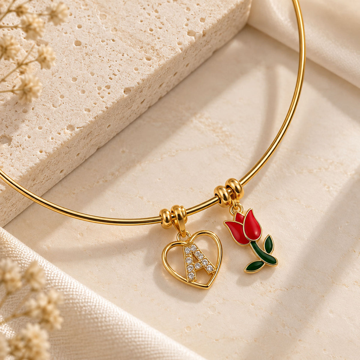 Gold-tone necklace arranged on a beige surface, featuring a heart-shaped initial A pendant with crystal detailing and a red floral charm on a delicate chain.