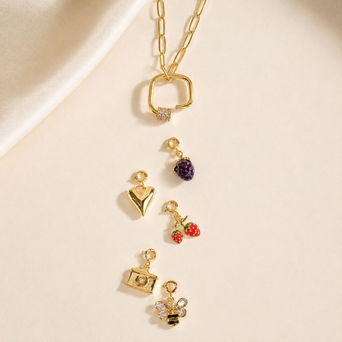 Gold-tone charm necklace with a detachable charm holder, featuring interchangeable purple bead, heart, strawberry, camera, and bee charms, displayed on a soft beige background.