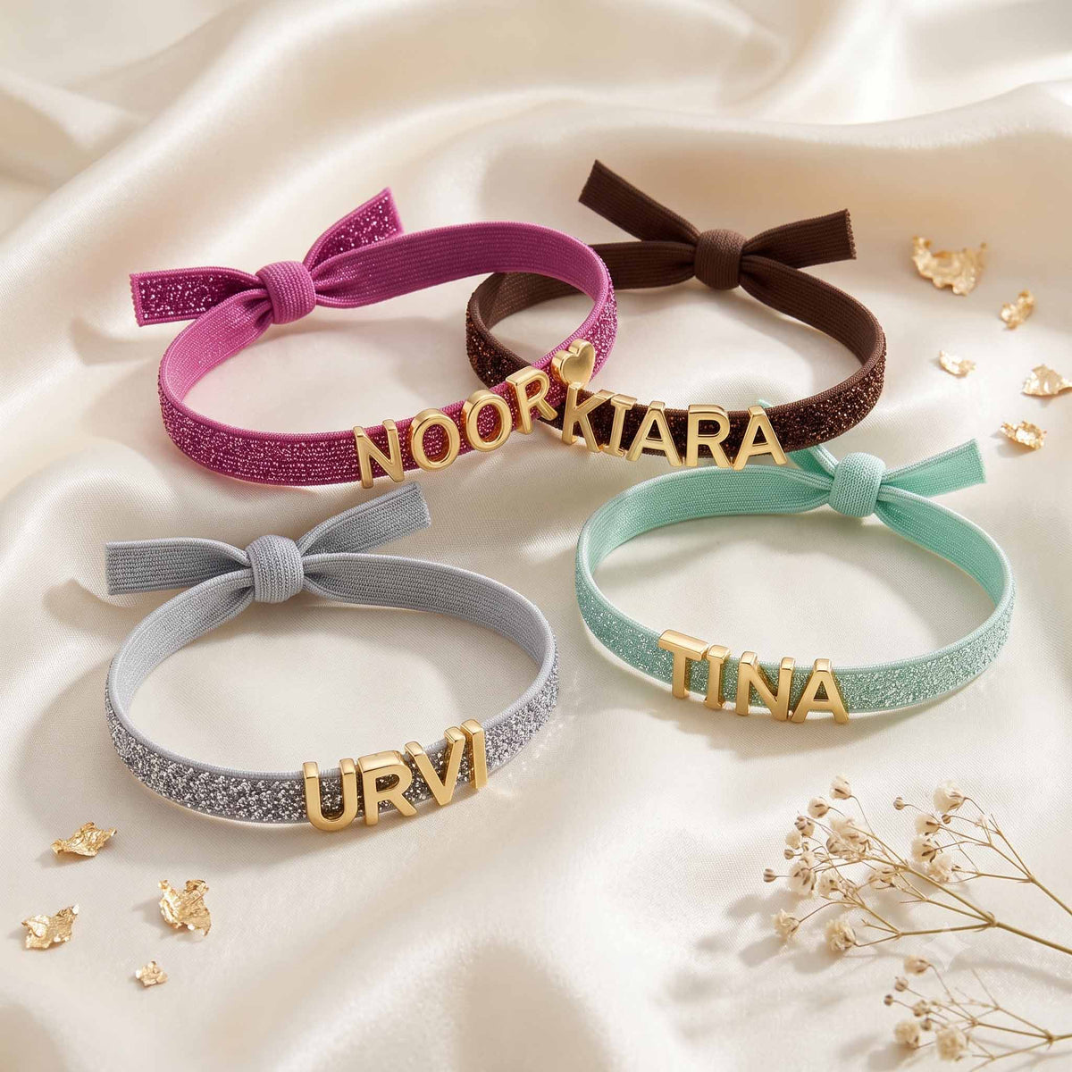 Lifestyle product image featuring four personalized glittery bracelets in pink NOOR, brown KIARA, mint green TINA, and grey URVI, arranged on white satin fabric with delicate dried flowers and gold decorative accents.