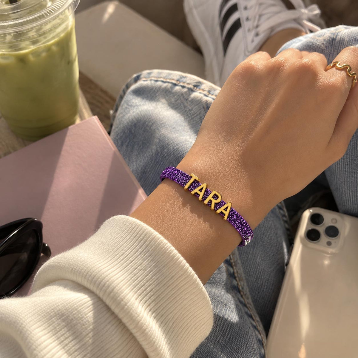 Lifestyle close-up of a hand wearing a purple glittery personalized bracelet with gold-tone TARA letter charms, styled with casual denim and a white sweatshirt.