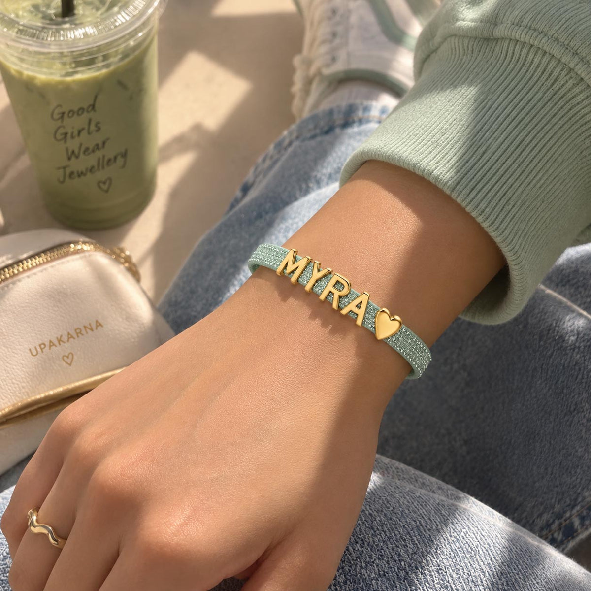 Lifestyle close-up of a hand wearing a mint-green glittery personalized bracelet with gold-tone MYRA letter charms and a heart charm, styled with a mint sweatshirt and denim.