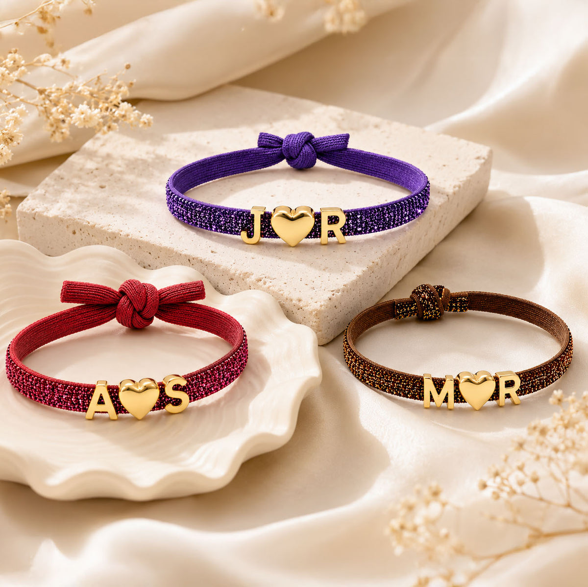 Three personalized glitter bracelets in purple, red, and brown, styled on cream-toned ceramic dishes and a stone pedestal, featuring gold letter-and-heart charms: J♥R, A♥S, and M♥R.