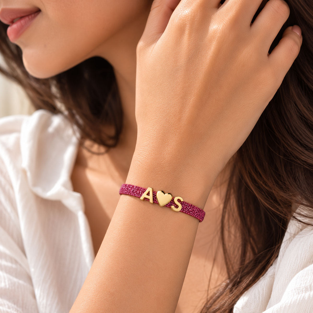 Pink glitter bracelet worn on a model’s wrist, featuring gold A♥S charms, styled with a white outfit in warm natural sunlight.