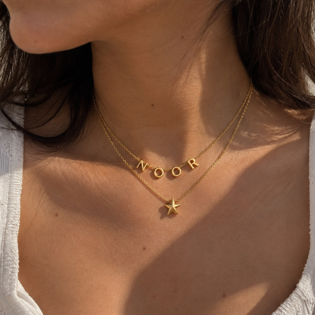 Close-up of a person wearing a stacked pair of delicate gold chain necklaces against a white top; the upper chain spells the name "NOOR" with smooth gold letter charms, while the lower chain holds a single dimensional star pendant.