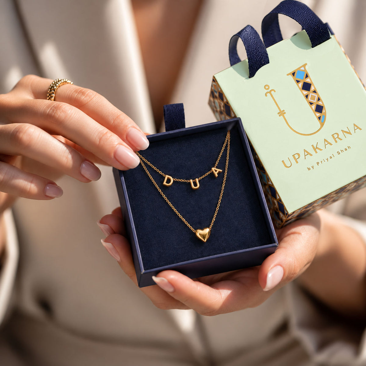 Close-up of hands holding an open dark blue jewelry box lined with velvet, displaying a layered gold chain necklace set with smooth letter charms spelling "DUA" and a solid gold heart pendant. Next to the box sits a miniature light green gift bag with blue ribbon handles and the brand name "UPAKARNA" printed on it.