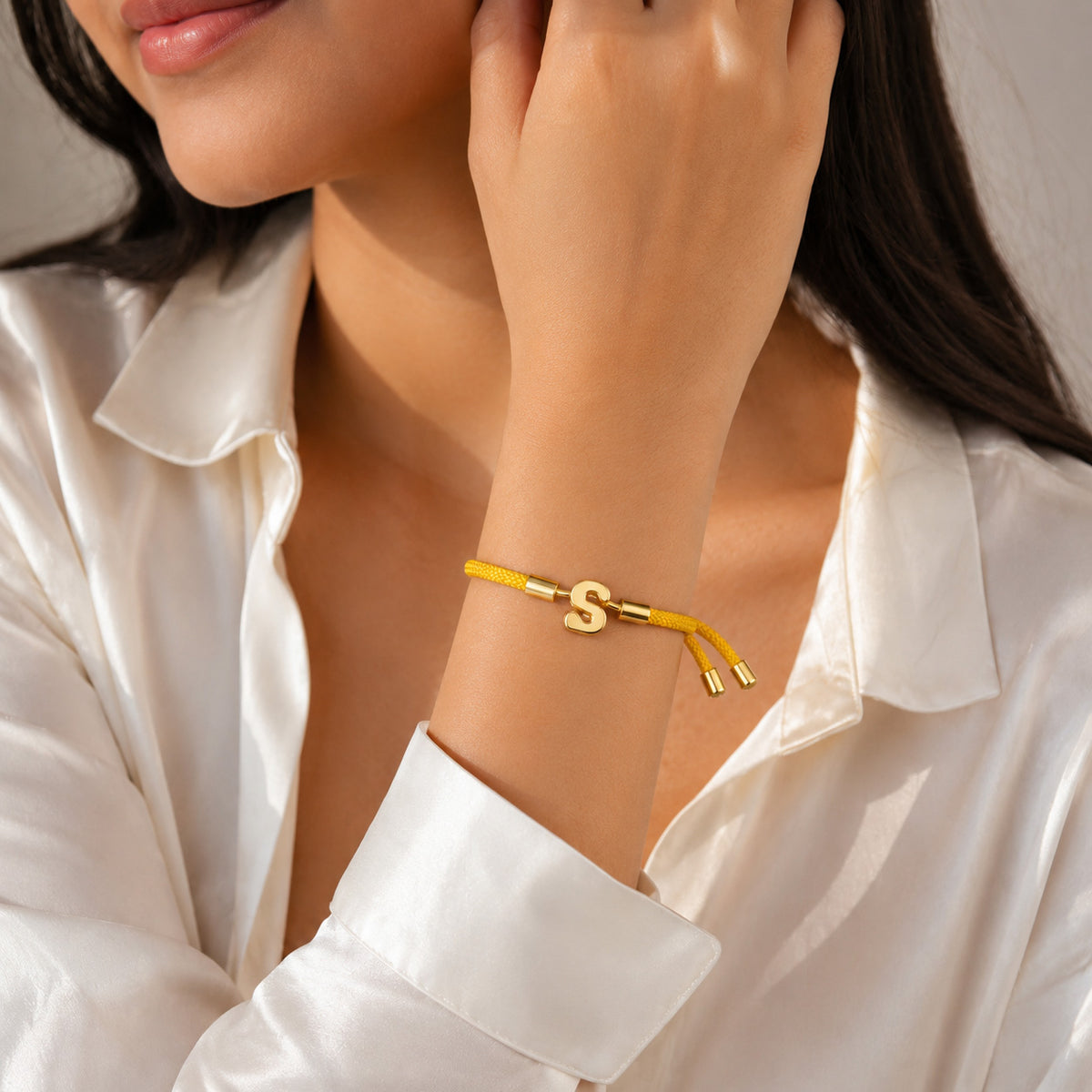 Gold-tone initial “S” charm bracelet with a yellow braided cord, worn on the wrist with a white satin shirt.