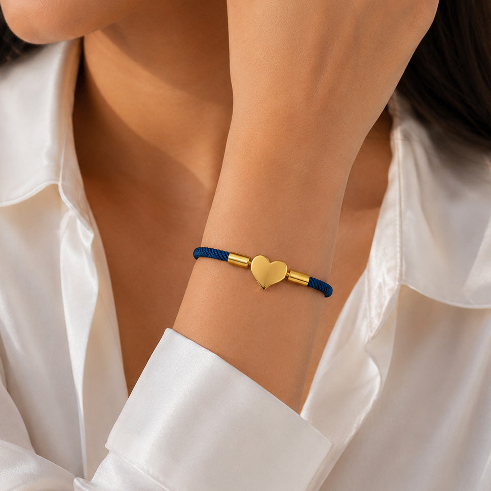 Gold-tone Heart charm bracelet with a blue braided cord, worn on the wrist with a white satin shirt.