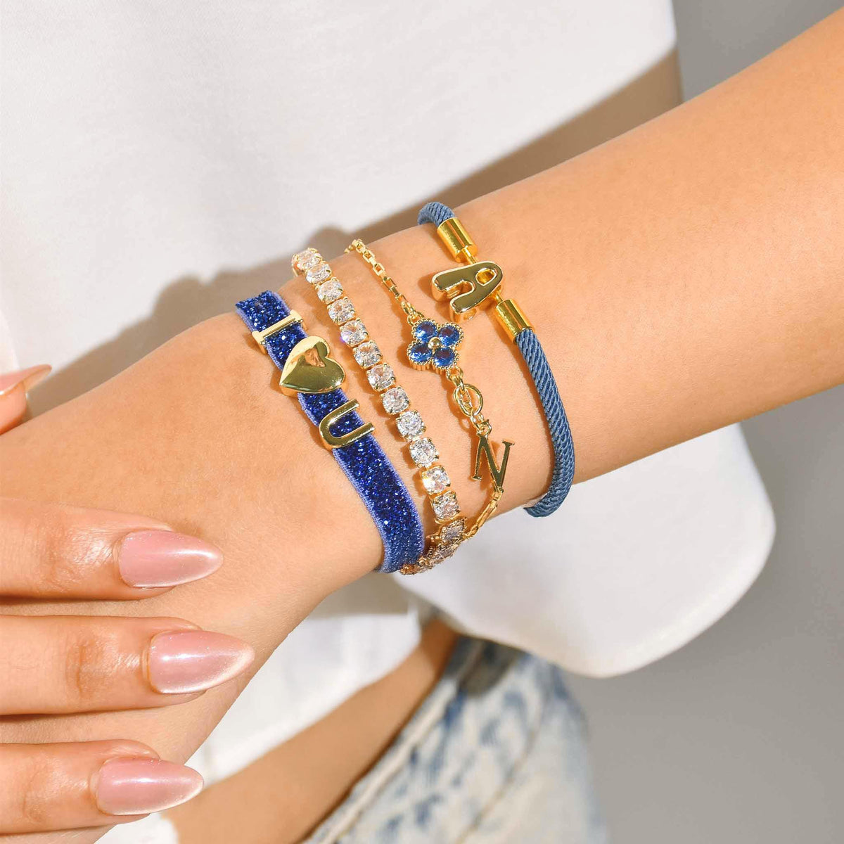 Close-up of a wrist wearing a layered stack of blue and gold bracelets, including a blue beaded bracelet with a heart charm, a sparkling tennis bracelet, a blue floral charm bracelet, and gold initial charms.