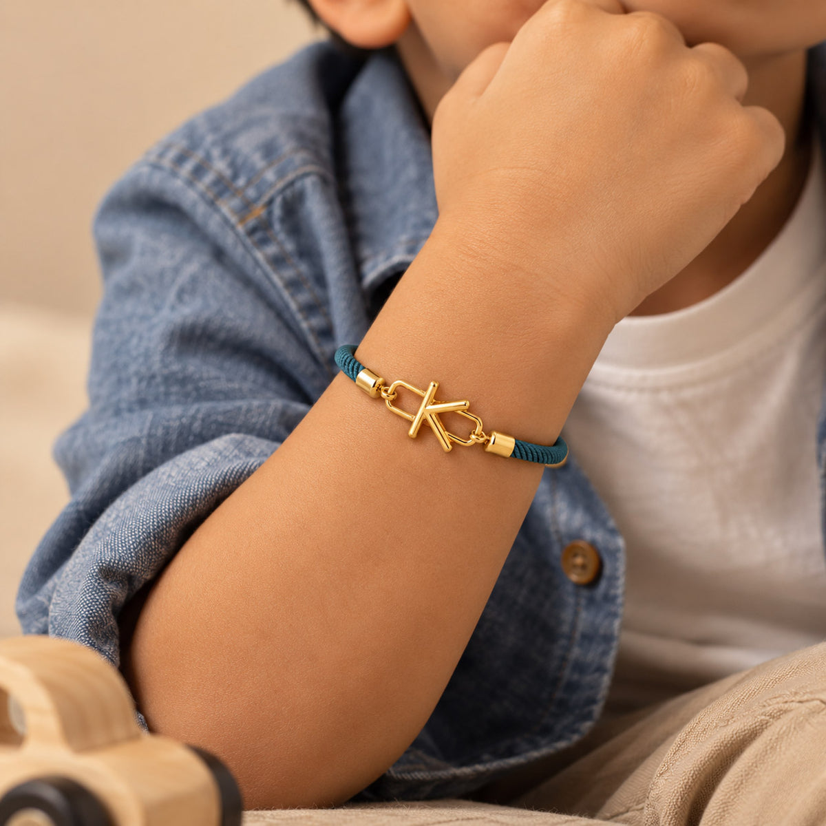 Close-up of a child wearing a teal braided cord bracelet with a gold-tone letter K charm, paired with a denim shirt and white top.