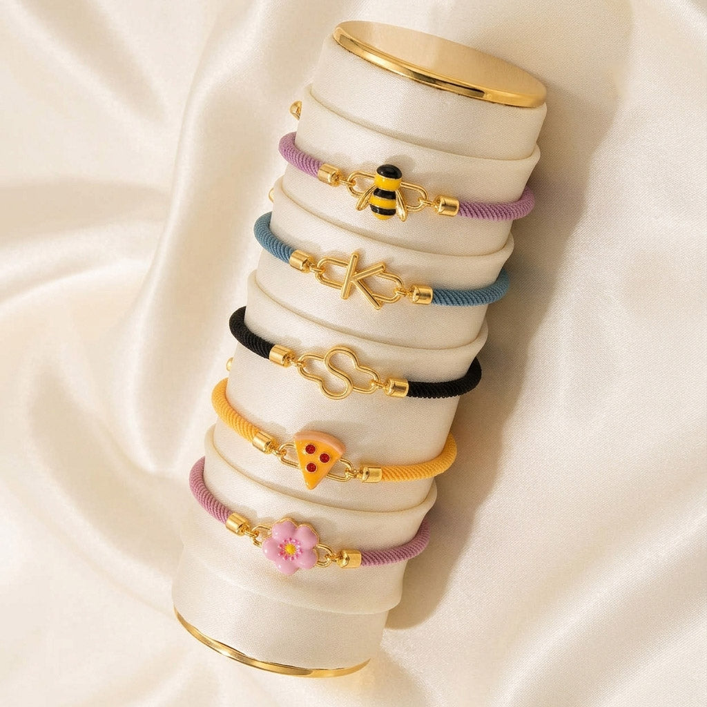 Six colorful braided cord bracelets with gold-tone charms, including bee, letter K, letter S, pizza, and pink flower designs, displayed on a white bracelet stand over cream satin fabric.