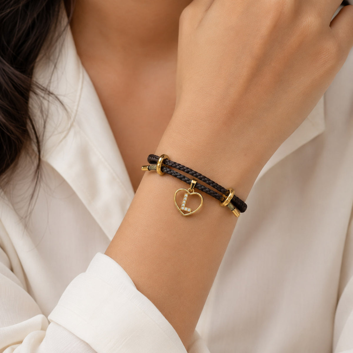 Close-up of a dark brown braided adjustable bracelet worn on the wrist, featuring a gold heart-shaped pendant with a sparkling letter L initial.