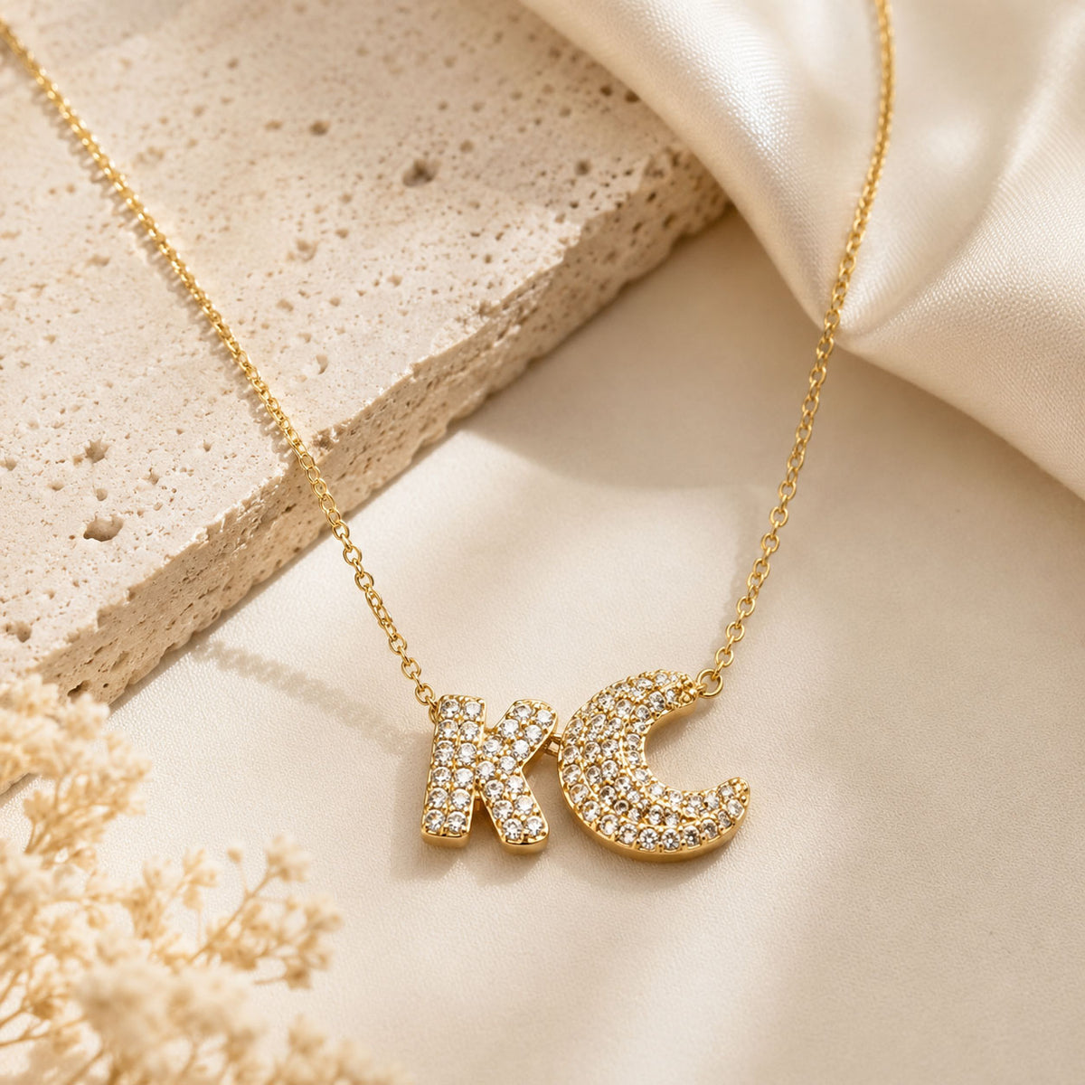 Gold-plated personalized necklace featuring a sparkling pavé “K” initial with a crescent moon charm, displayed on elegant ivory satin with a premium, warm editorial aesthetic