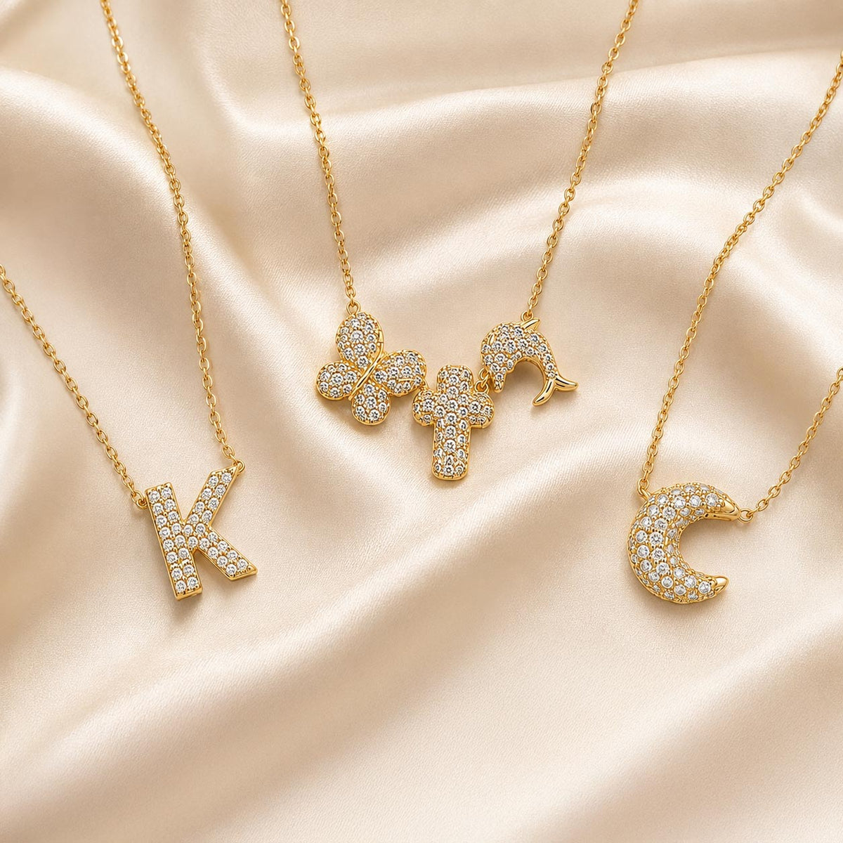 Gold-plated necklace collection featuring a pavé K initial, butterfly & cross charm necklace, and crescent moon necklace, arranged on luxurious ivory satin.