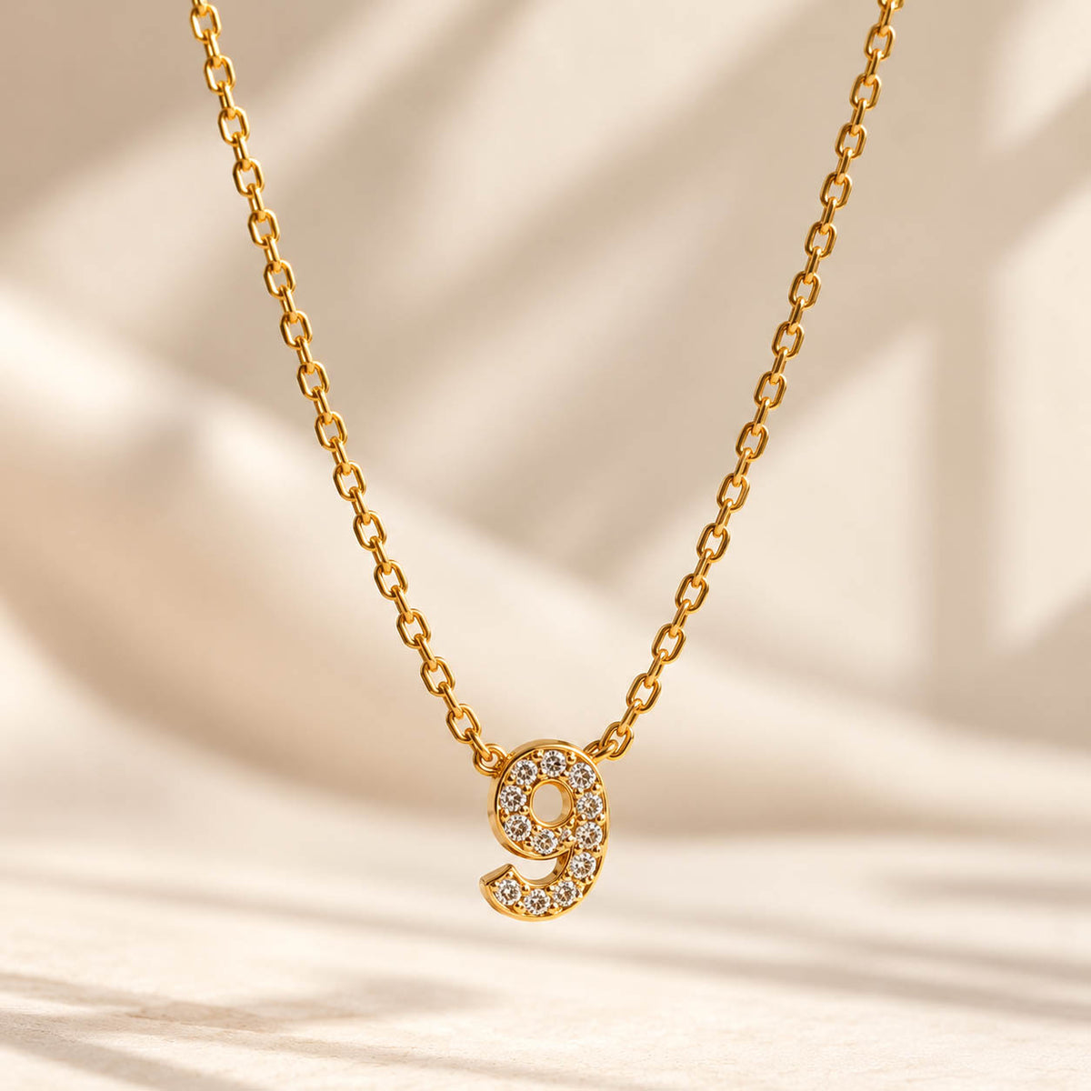A studio shot of a thin gold link necklace centered with a small number "9" charm entirely encrusted with sparkling rhinestones, laid out on a light surface with soft lighting.