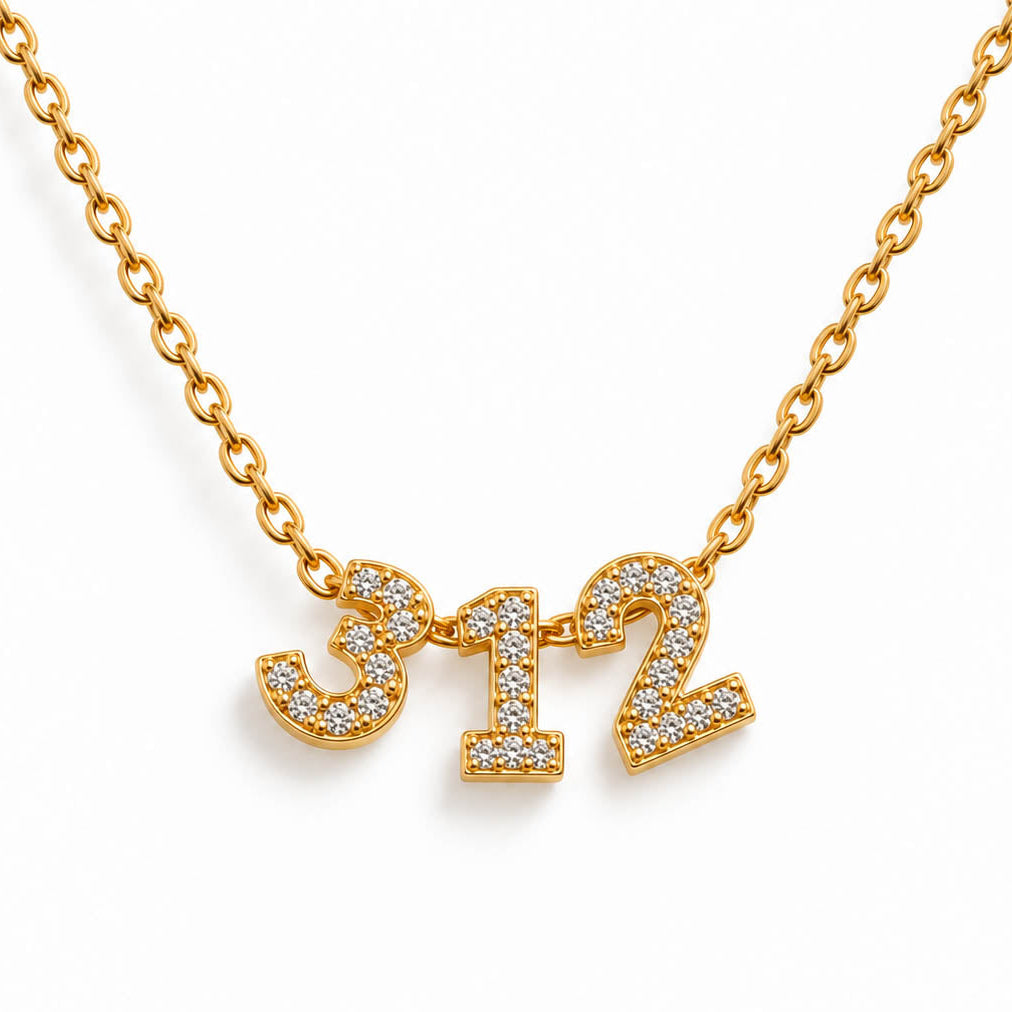A studio shot of a thin gold link necklace centered with three rhinestone-encrusted charms that spell the numbers "3", "1", and "2".