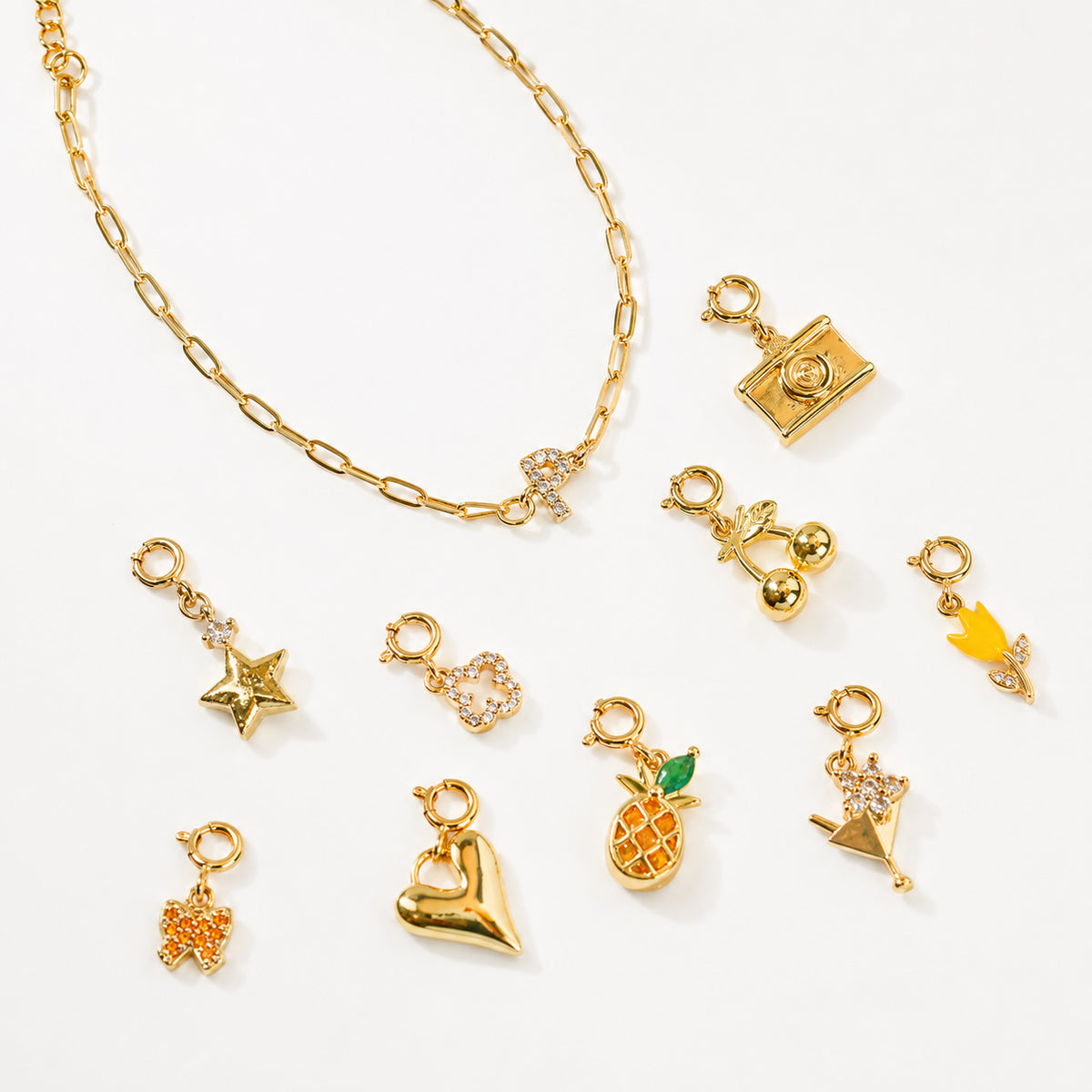 Gold-tone charm bracelet with a sparkling letter R charm, displayed with interchangeable charms including a camera, musical notes, star, flower, pineapple, crown, butterfly, heart, and hand-shaped charm on a white background.