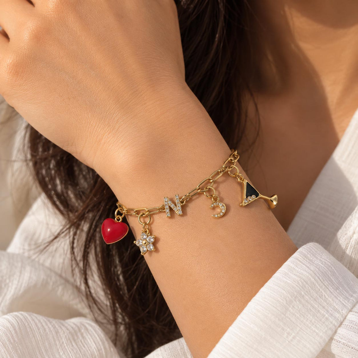 Gold-tone charm bracelet worn on a wrist, featuring a red heart, sparkling letter N, flower charm, crescent moon, and martini glass charm, styled with a white outfit.