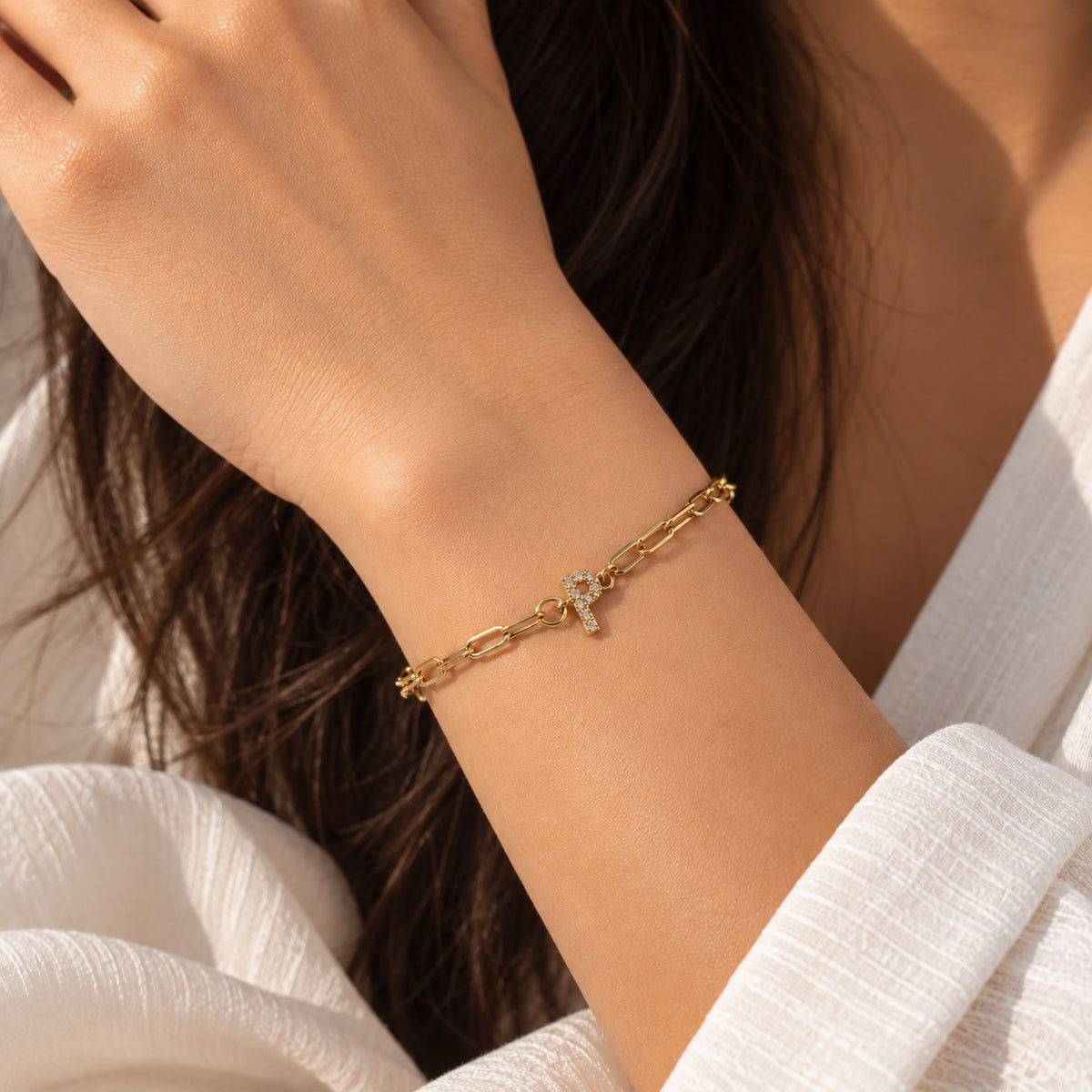 Gold-tone chain bracelet worn on a wrist, featuring a small sparkling letter P charm, styled with a white outfit in warm natural light.