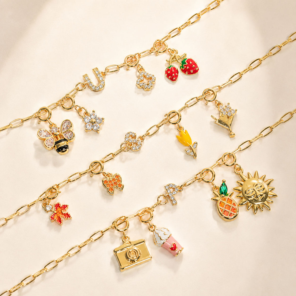 Gold-tone charm chain bracelets arranged on a cream background, featuring colorful charms including strawberries, a bee, butterfly, crown, flower, star, pineapple, sun, camera, and ice cream.