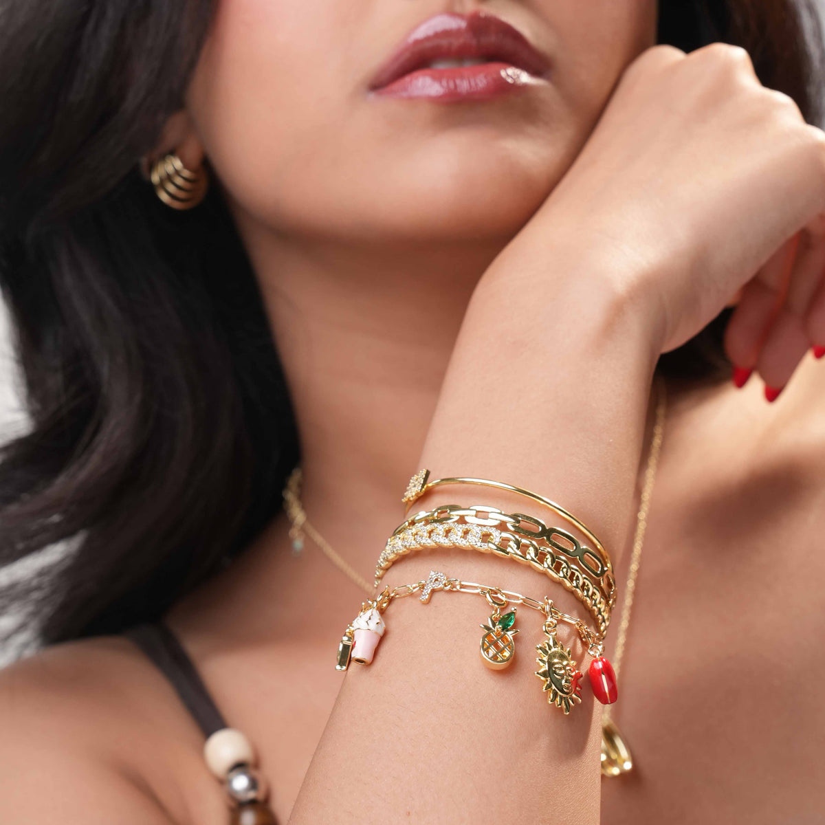 Close-up of a woman wearing layered gold-tone bracelets, including a charm bracelet with pineapple, sun, red charm, and drink charms, styled with gold bangles and chain bracelets.
