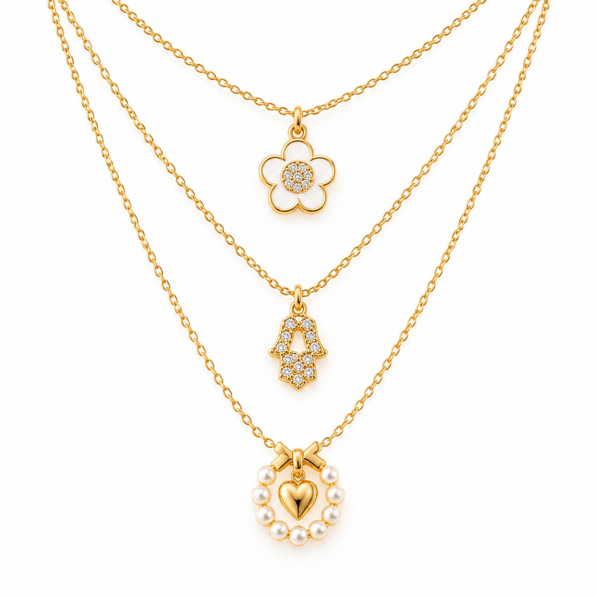 A product photo of a layered gold-tone necklace set featuring a daisy flower pendant with a white enamel outline and crystal center, a rhinestone-paved Hamsa hand charm, and a pearl wreath pendant holding a glossy gold heart beneath a bow.