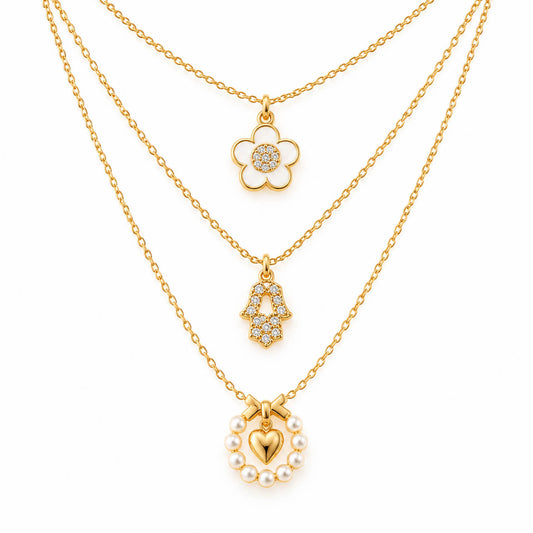 A product photo of a layered gold-tone necklace set featuring a daisy flower pendant with a white enamel outline and crystal center, a rhinestone-paved Hamsa hand charm, and a pearl wreath pendant holding a glossy gold heart beneath a bow.