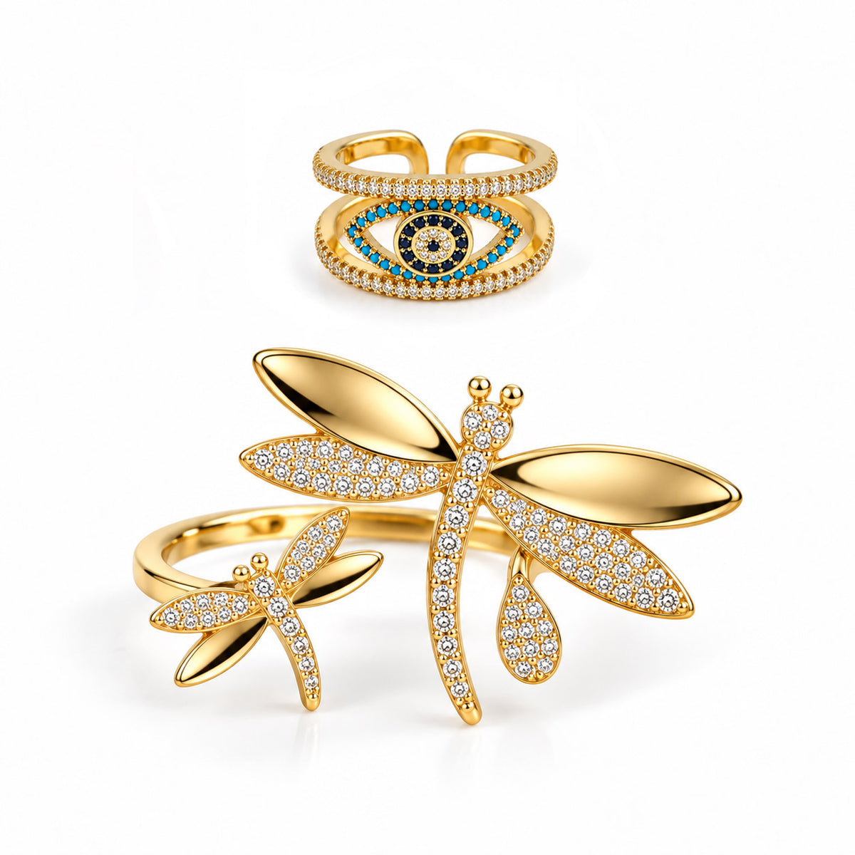 A product photo of two gold-tone rings featuring an open band with a prominent evil eye motif framed by crystal-paved rows and turquoise accents, and an open wrap-around ring showcasing large crystal-paved dragonfly charms with polished gold wings