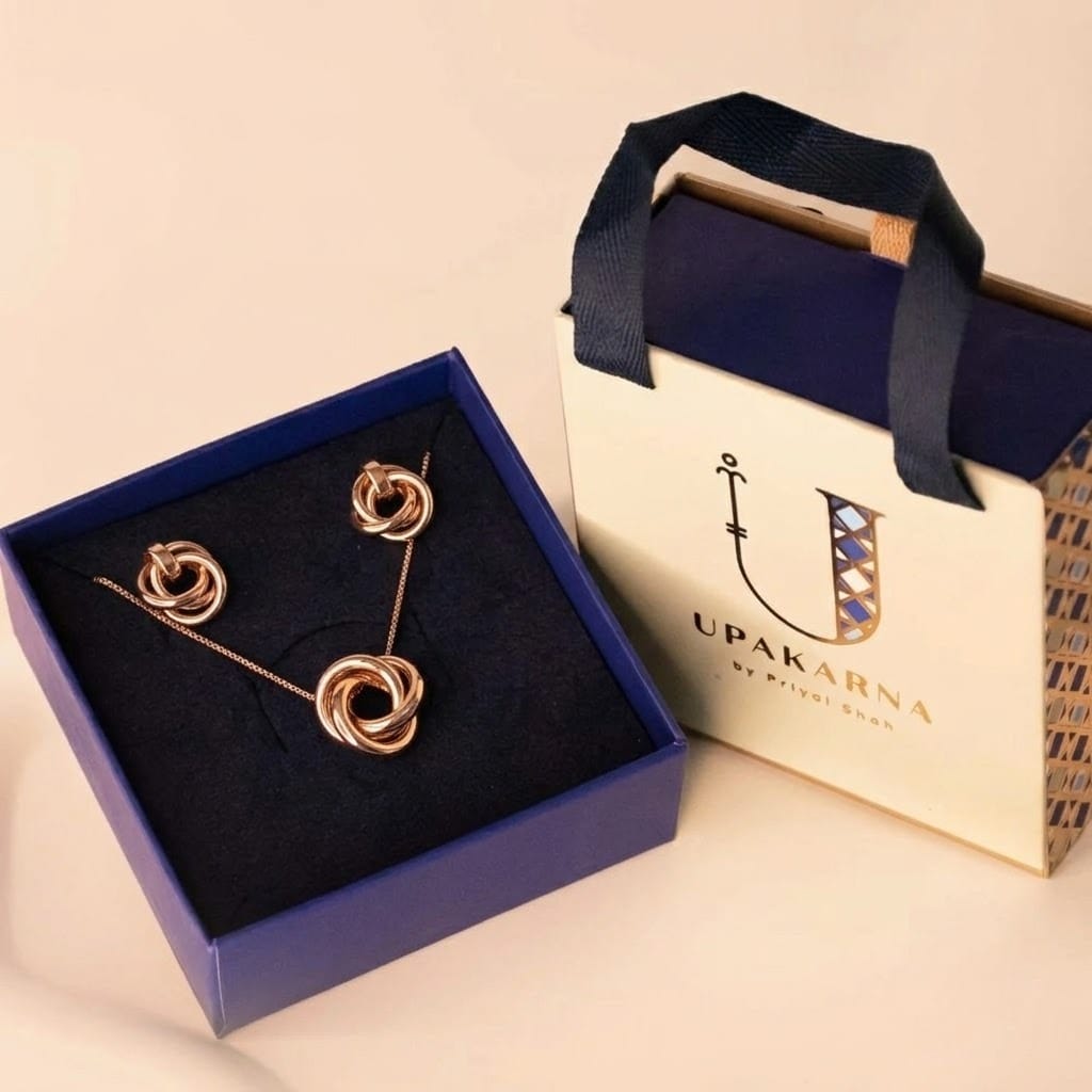 Luxury rose gold jewellery gift set featuring a pendant necklace and matching knot stud earrings displayed in a premium navy blue jewellery box. The set is presented alongside an elegant Upakarna branded gift bag with navy ribbon handles, making it ideal for gifting, special occasions, and celebrations.