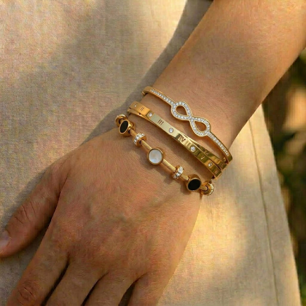 Luxury gold anti-tarnish bracelet stack featuring a crystal infinity bangle, Roman numeral bangle, and a mother-of-pearl cable bracelet with sparkling crystal accents, elegantly worn on the wrist in natural sunlight.
