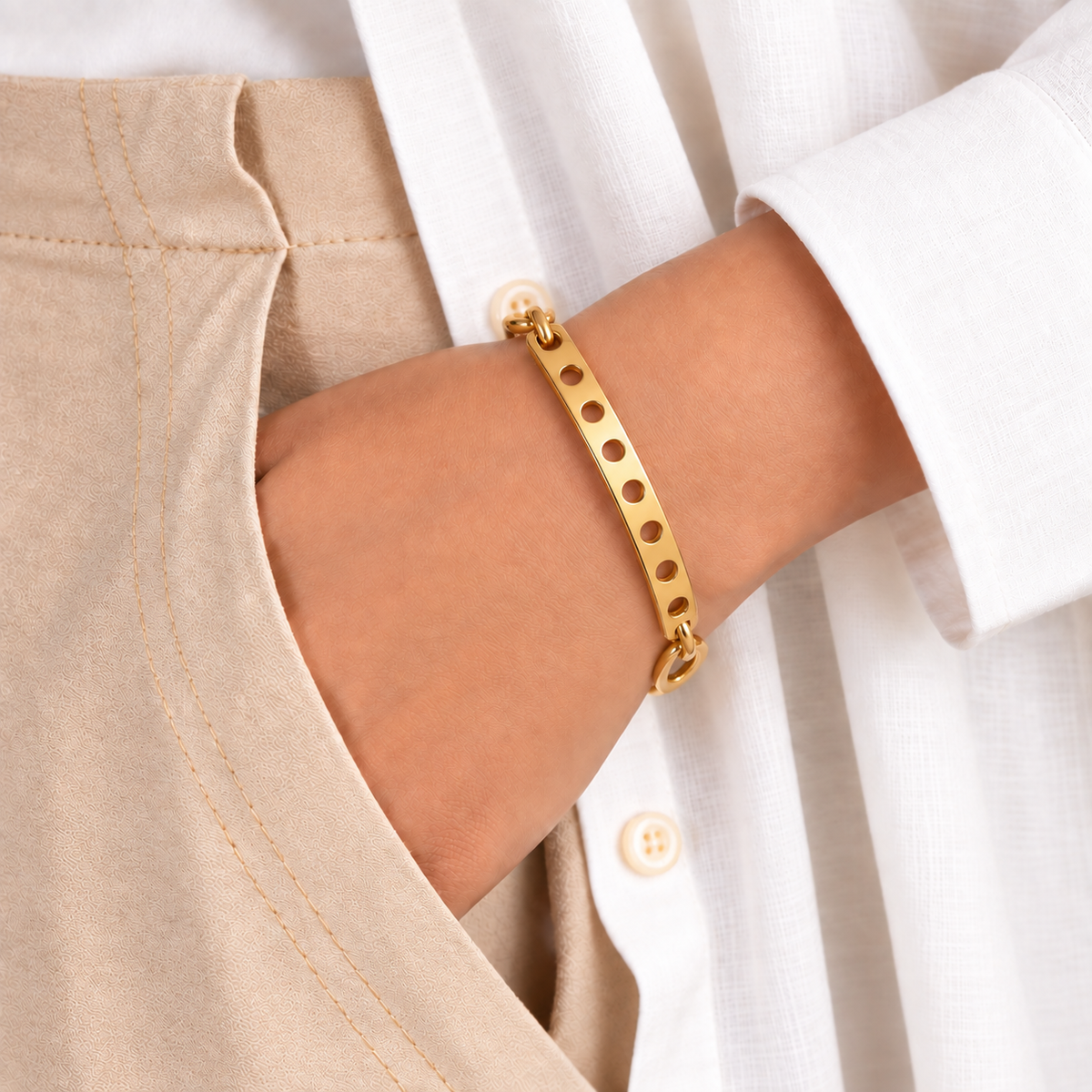 Gold-tone bangle bracelet with seven circular cutouts worn on a wrist with a white shirt and beige fabric.