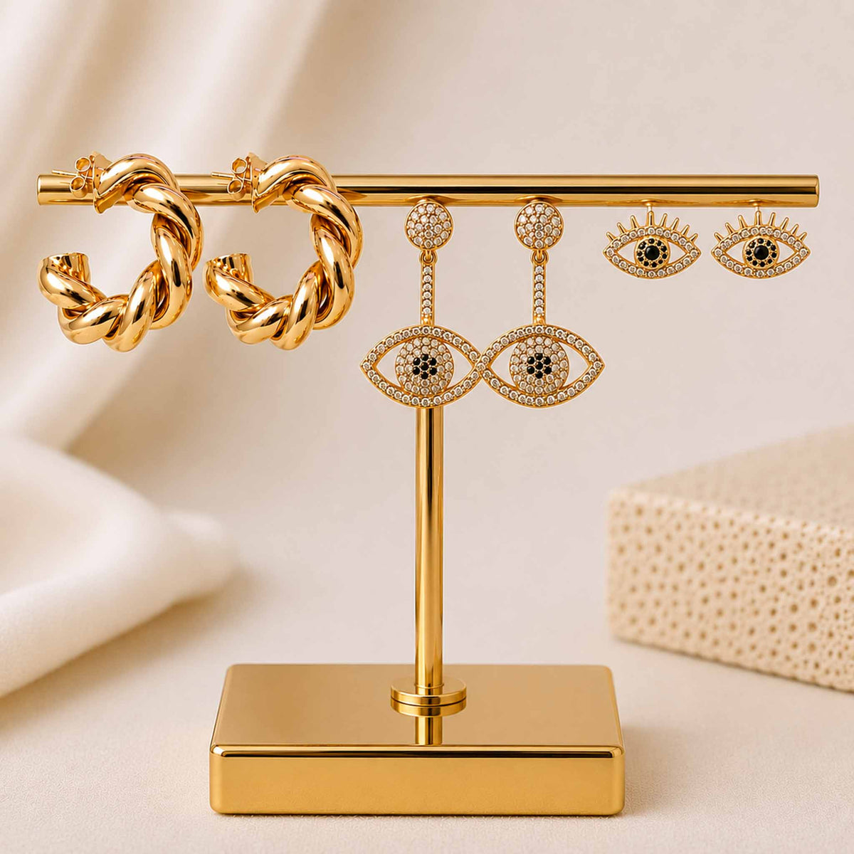 Gold-tone earring stack featuring twisted hoops, sparkling evil eye studs, and celestial eye dangler earrings displayed on a gold earring stand against a soft beige background.