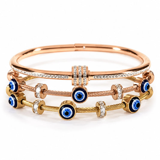product photo of a stacked set of rose gold and gold-tone bracelets featuring a crystal-accented bangle with a triple-paved barrel spacer, paired with twisted cable wire bands decorated with blue-and-white evil eye charms and rhinestone spacers.