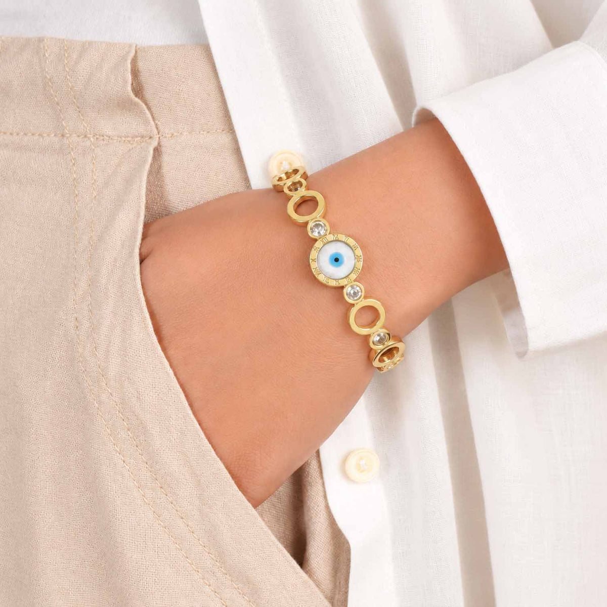 Gold link bracelet with a central blue evil eye and Roman numeral design, polished oval links, and crystal accents, worn on the wrist with a neutral beige and white outfit.