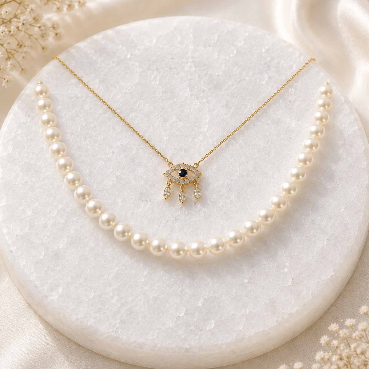 Gold evil eye pendant necklace paired with a pearl strand necklace displayed on a white circular jewellery stand.
