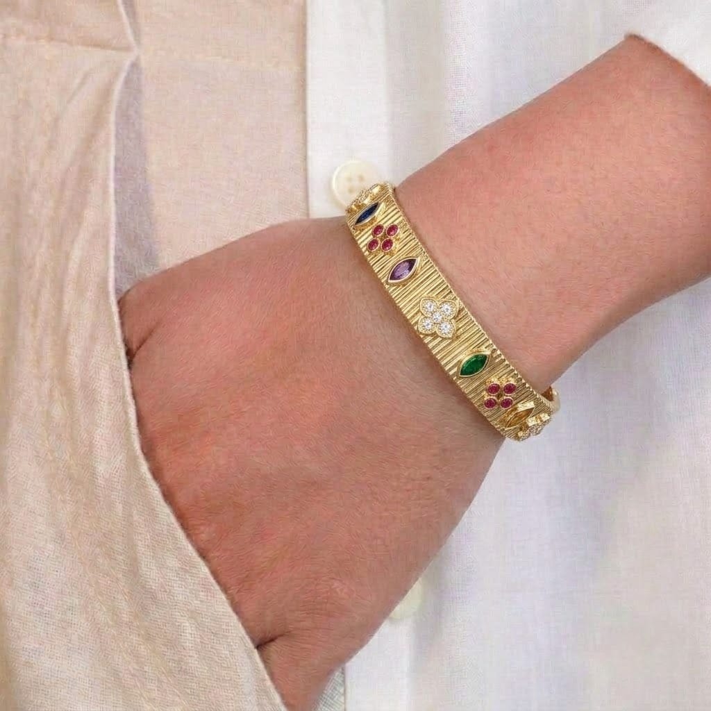 Close-up of a hand wearing an elegant gold-tone open cuff bangle with a textured ribbed finish, featuring colorful marquise and round gemstone accents and a sparkling crystal floral centerpiece. The anti-tarnish statement bracelet is styled with a neutral long-sleeve outfit, showcasing its vibrant design for everyday wear, festive occasions, and elegant wrist stacking.