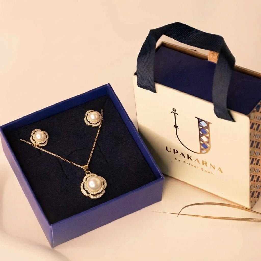A gold-tone jewelry set featuring a delicate chain necklace with a floral pendant centered around a pearl, surrounded by a textured scalloped design. The set includes matching stud earrings with the same pearl floral motif, displayed inside an open navy blue box. Beside it is an elegant Upakarna branded gift bag in cream and navy with ribbon handles.